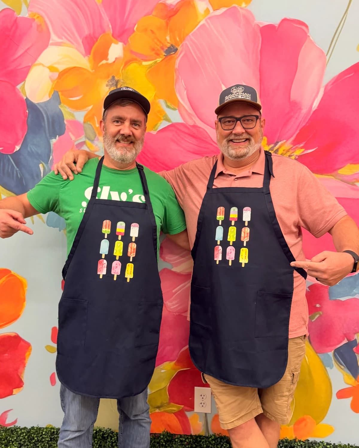 Savannah Square Pops founders Mark and Judd in pop-print aprons in front of a flower mural