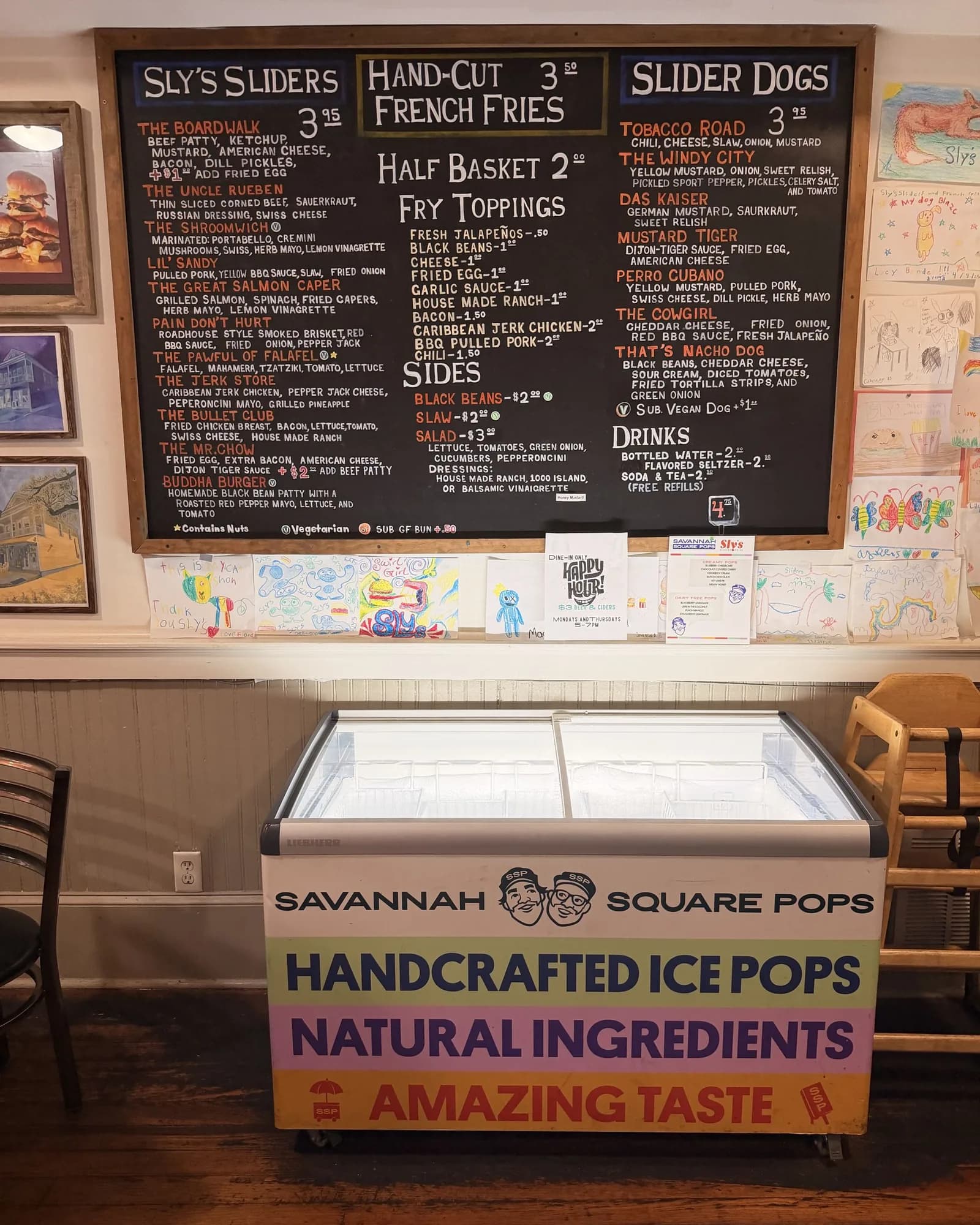 A branded Savannah Square Pops freezer on the floor at Sly's Sliders, beneath the shop's chalkboard menu